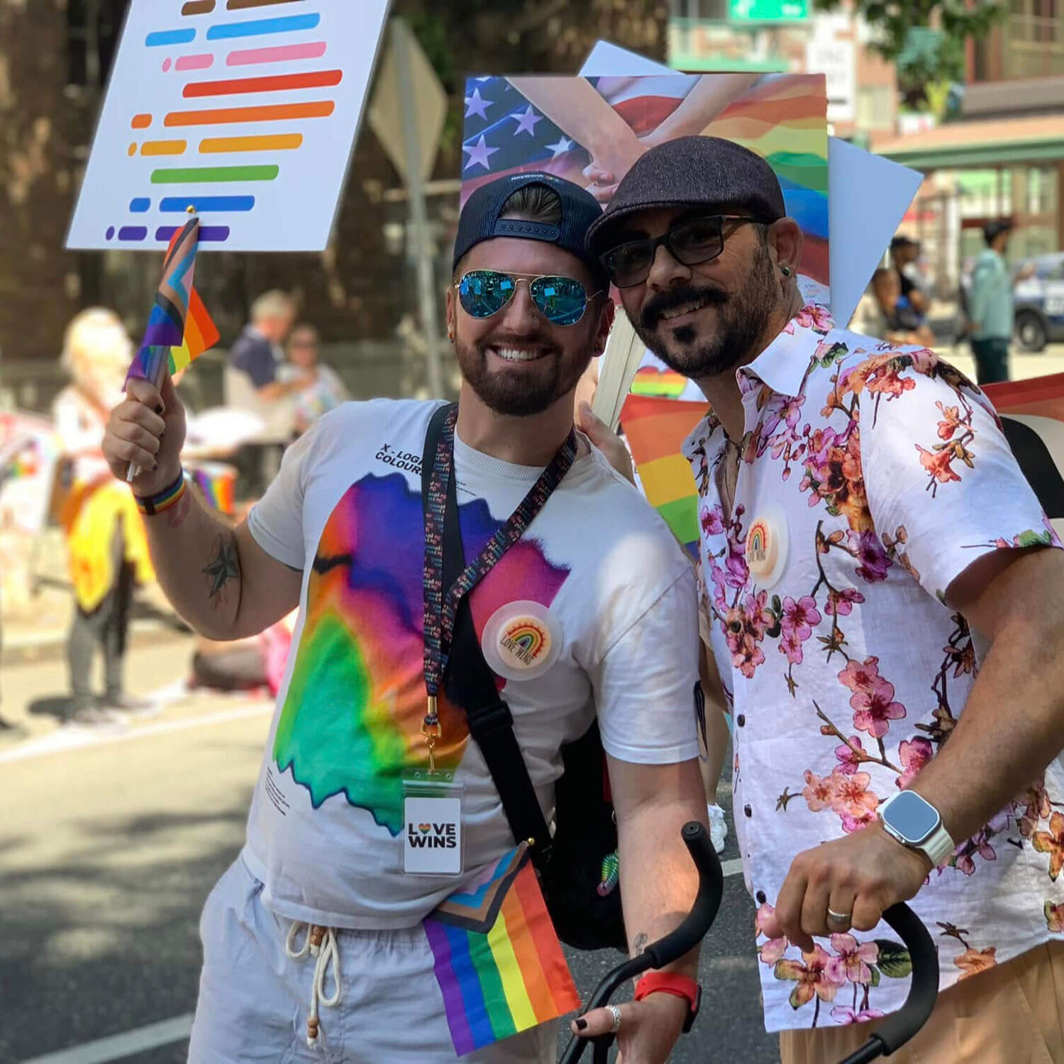 485138421_1063225655851339_8397134104894188940_n Two people smiling and posing together at a Pride parade, holding small rainbow flags